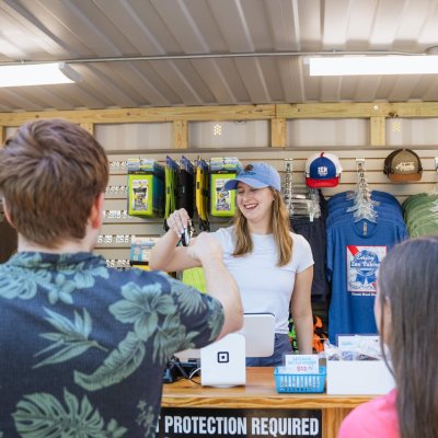 Two customers at a merchandise stand with clothing and hats, served by a smiling cashier.