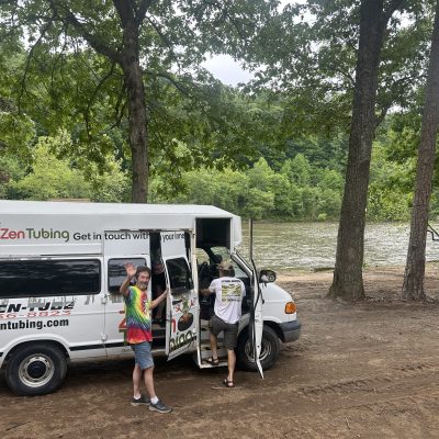 Man waves beside van with 'Zen Tubing' logo, parked by a river and trees.